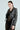 A person with slicked-back hair wears the DEMI EMBELLISHED MOTO jacket over a sheer black lace dress, accessorized with hoop earrings, and confidently poses against a plain backdrop.