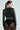 Seen from behind, a person with a ponytail wears the DEMI EMBELLISHED MOTO jacket paired with black lace leggings and large hoop earrings, all set against a light gray background. Their edgy style is highlighted by the bold design of the faux leather jacket.