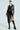 Wearing the DEMI EMBELLISHED MOTO over a black, sheer lace dress with a high slit and lace-up black platform boots, they stand confidently against a plain background.