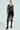 A person stands confidently in a black lace dress with a slit, layered under the DEMI EMBELLISHED MOTO jacket. They wear knee-high platform leather boots and large hoop earrings against a plain light background.