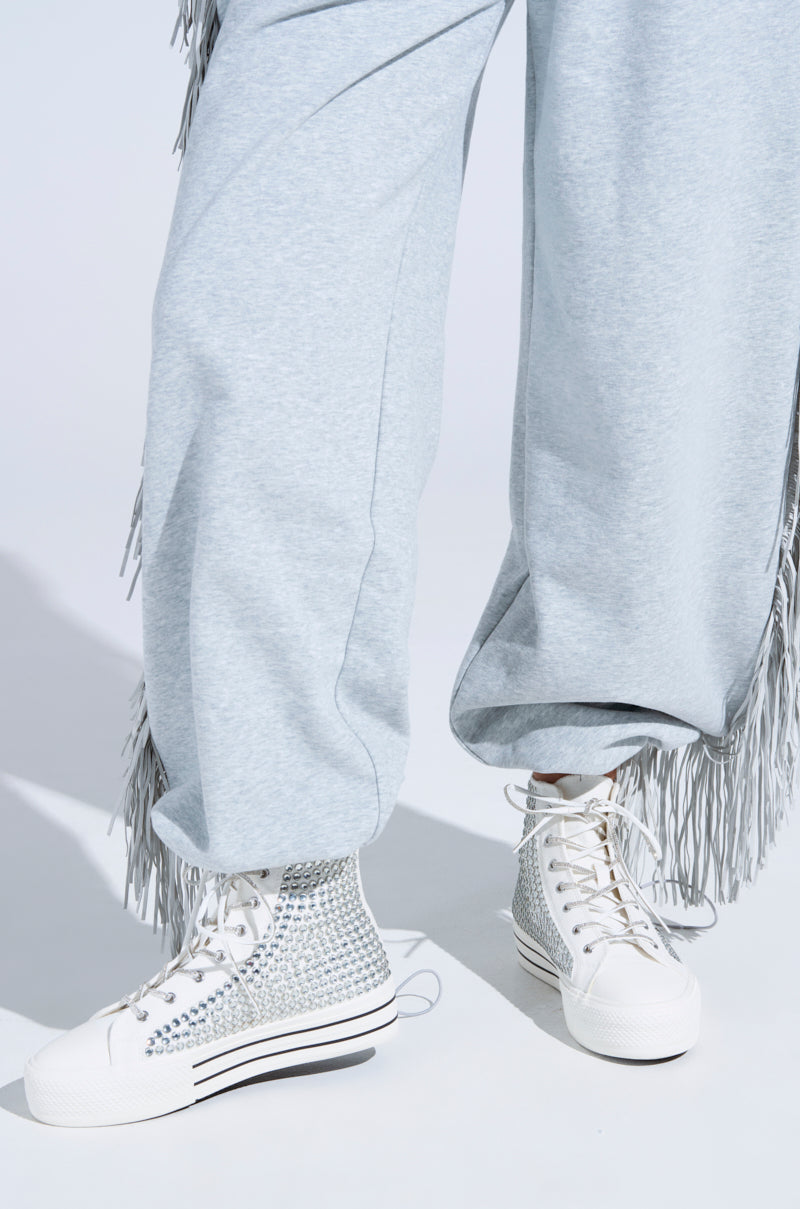 A person wearing light gray fringe pants and the AZALEA WANG ZAFRINA WHITE HIGH TOP SNEAKER with platform soles and silver studs stands on a white surface. Only their lower legs and feet are visible.