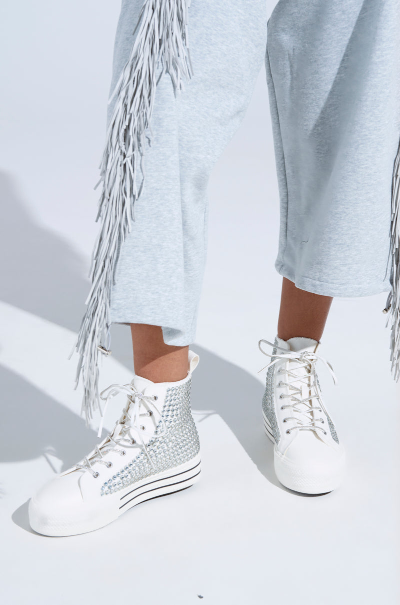 Wearing light gray, fringed pants and striking AZALEA WANG ZAFRINA WHITE HIGH TOP SNEAKERS with rhinestone details and thick platform soles, the person stands on a white surface.