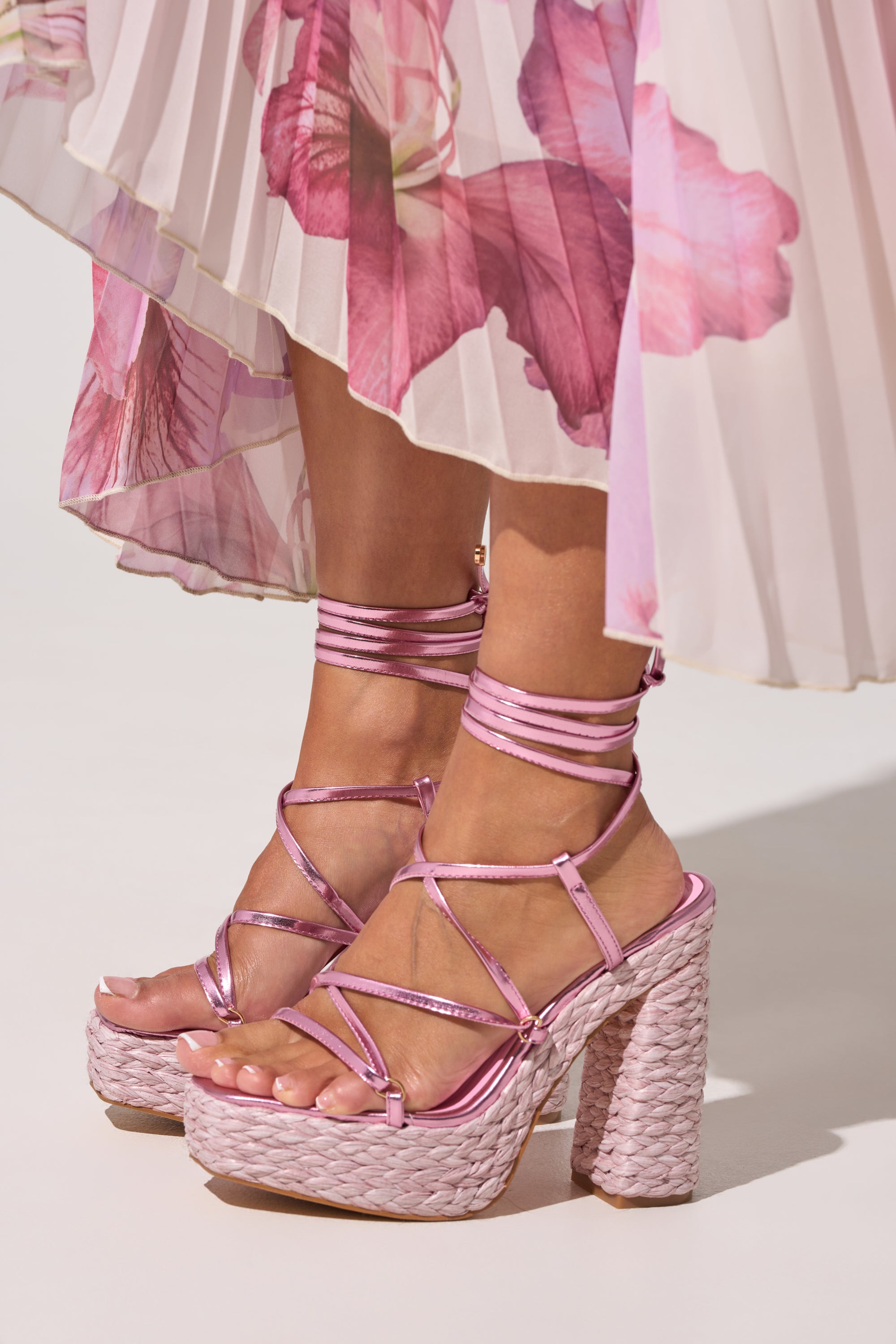Someone wearing the AKIRA GOLD ZAIDEE Pink Platform Raffia Lace Up Sandal, styled with a flowy pleated skirt adorned with large pink floral prints. 0