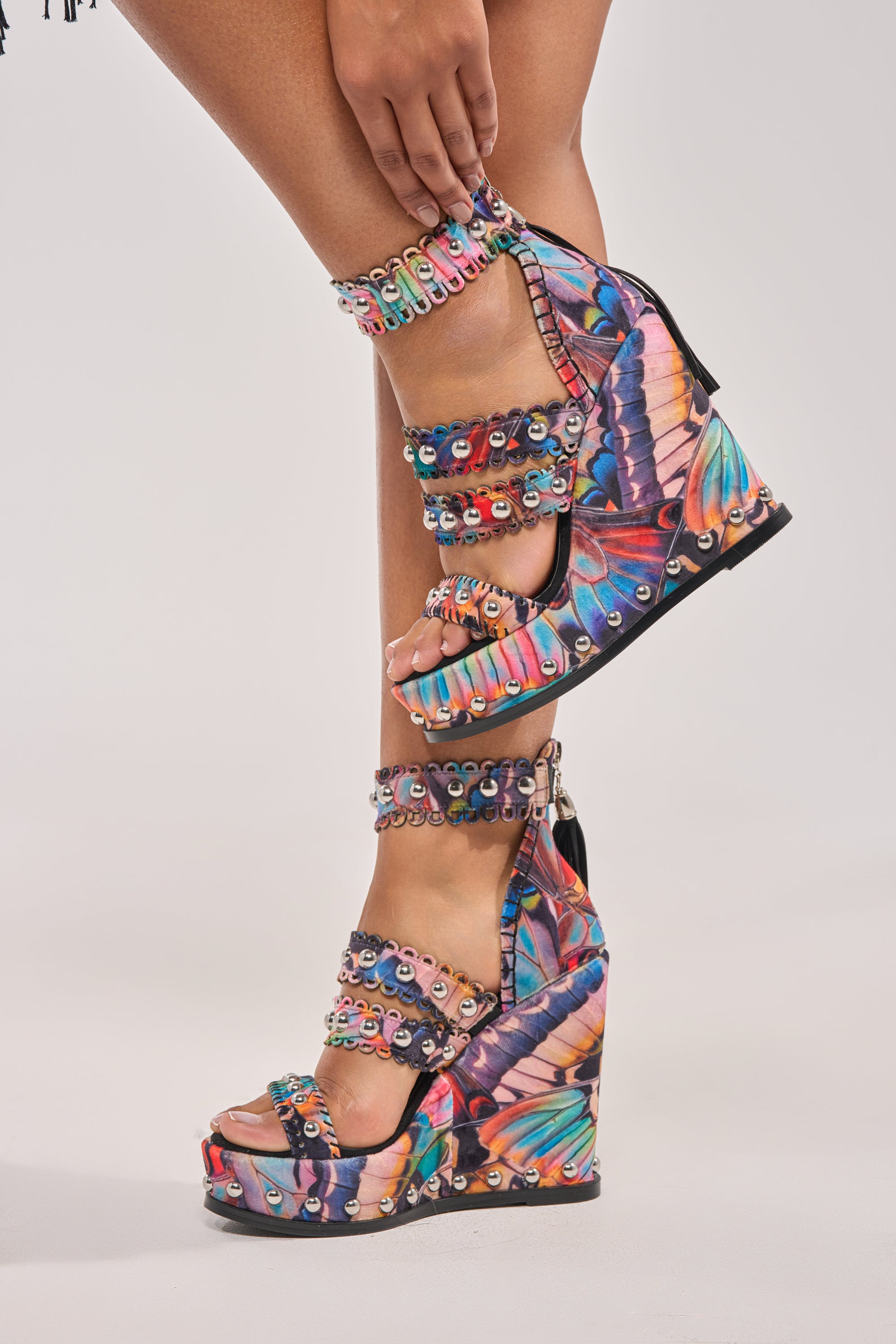 Close-up of a person standing on tiptoe in the AKIRA GOLD ZANIA BUTTERFLY MULTI PRINT FRINGE EMBELLISHED WEDGE SANDAL, featuring butterfly wing patterns, sequin ankle straps, a comfort cushion insole, and sky-high wedge heels. 1