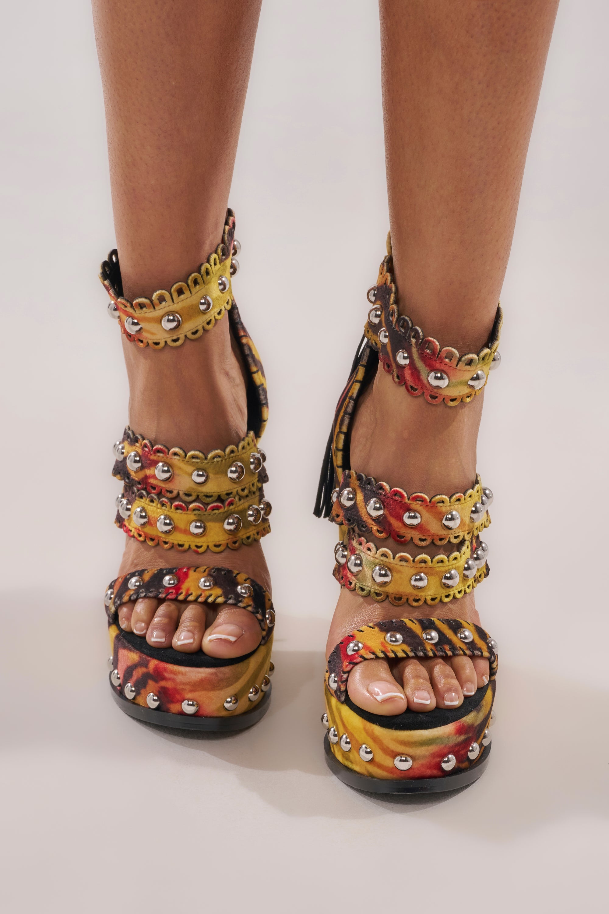 A pair of feet wearing AKIRA GOLD ZANIA TIGER MULTI PRINT FRINGE EMBELLISHED WEDGE SANDALS, featuring a bold wedge sole, multiple ankle and foot straps, fringe details, and vibrant multi-colored patterns. 1