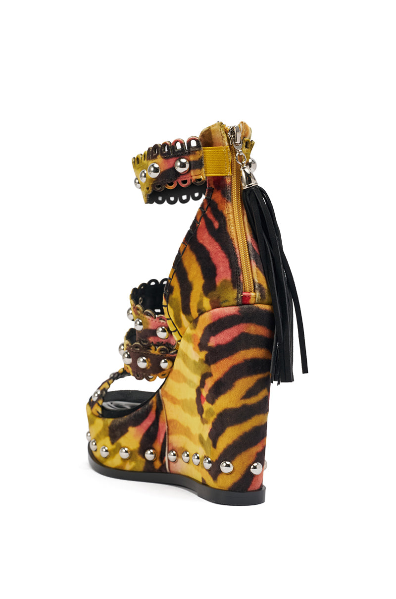 The AKIRA GOLD ZANIA TIGER MULTI PRINT FRINGE EMBELLISHED WEDGE SANDAL features a high wedge heel, bold tiger print in yellow, orange, and black, multiple studded straps with metal eyelet accents, and a back zipper with a black tassel. 6