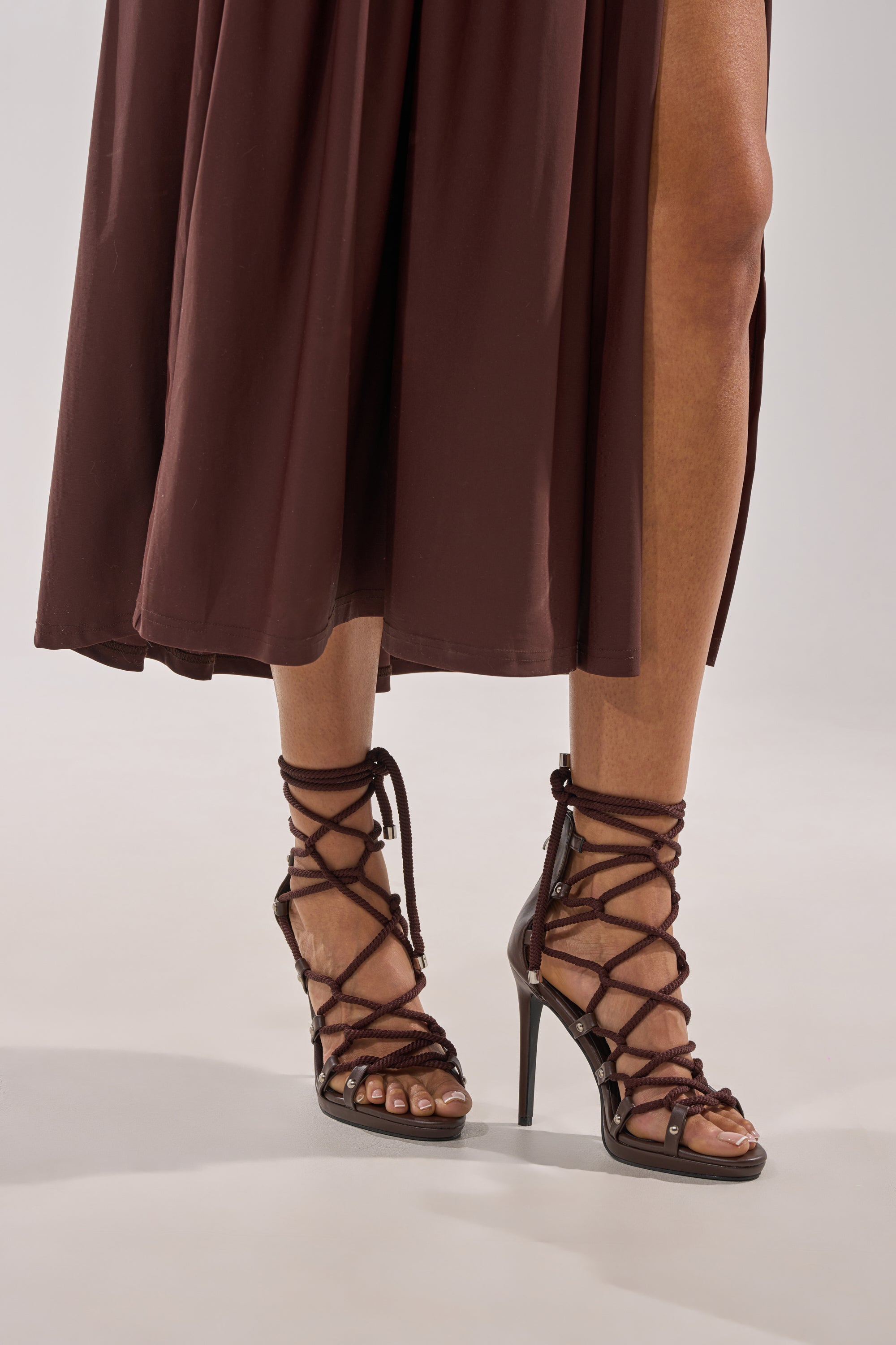A person wearing a midi brown skirt and the AZALEA WANG BROWN PU ROPE STRAP STILETTO SANDAL, featuring chocolate brown wrap-around rope laces and a faux leather stiletto heel elegantly crisscrossing the ankles. 2