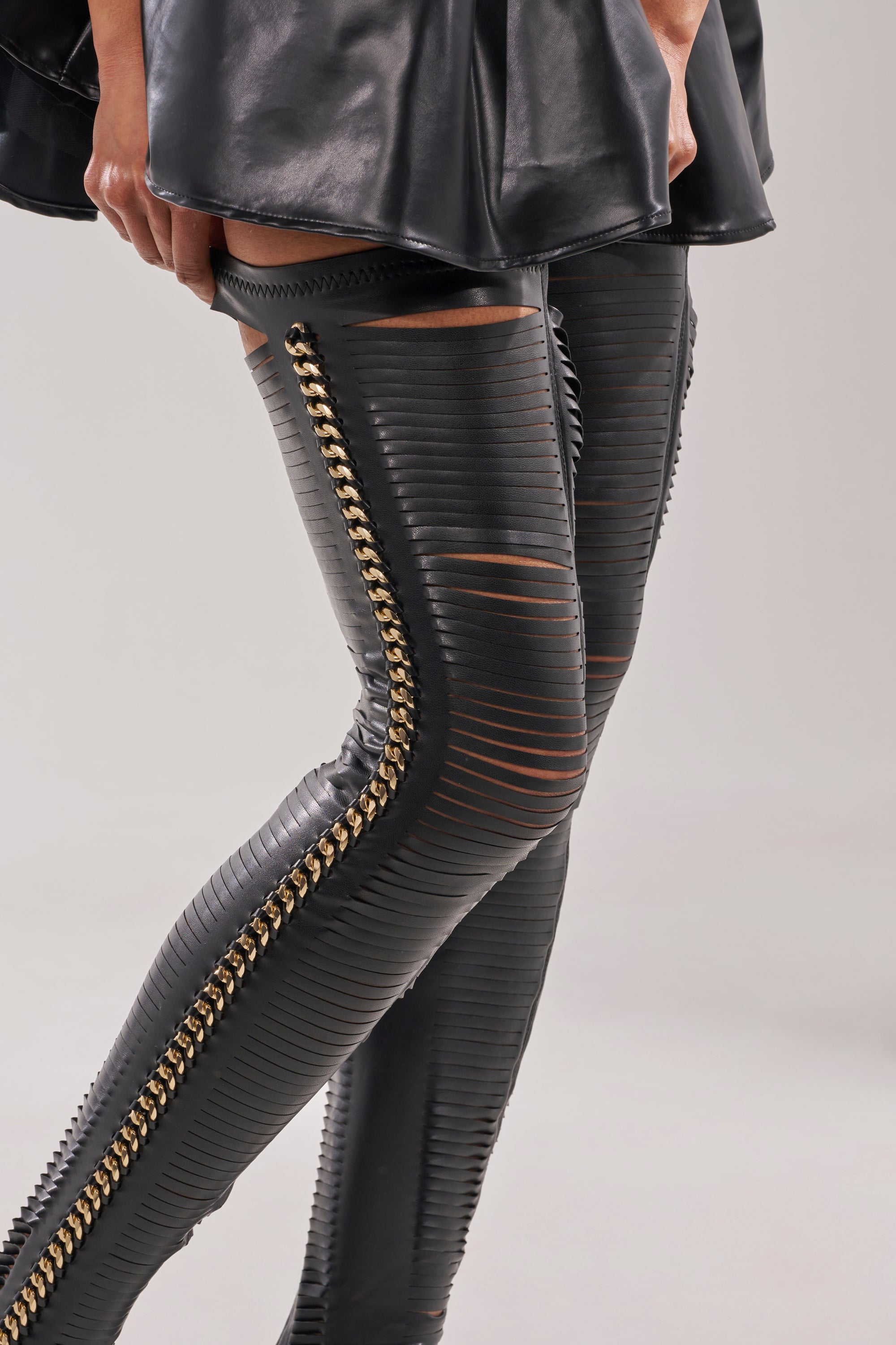 A person styles the AZALEA WANG ZARIE BLACK THIGH HIGH STRETCH PU GOLD EMBELLISHED STILETTO BOOT with fitted, black ribbed leather pants featuring chain and gold hardware details, plus a short black leather skirt and horizontal pant cutouts. 2