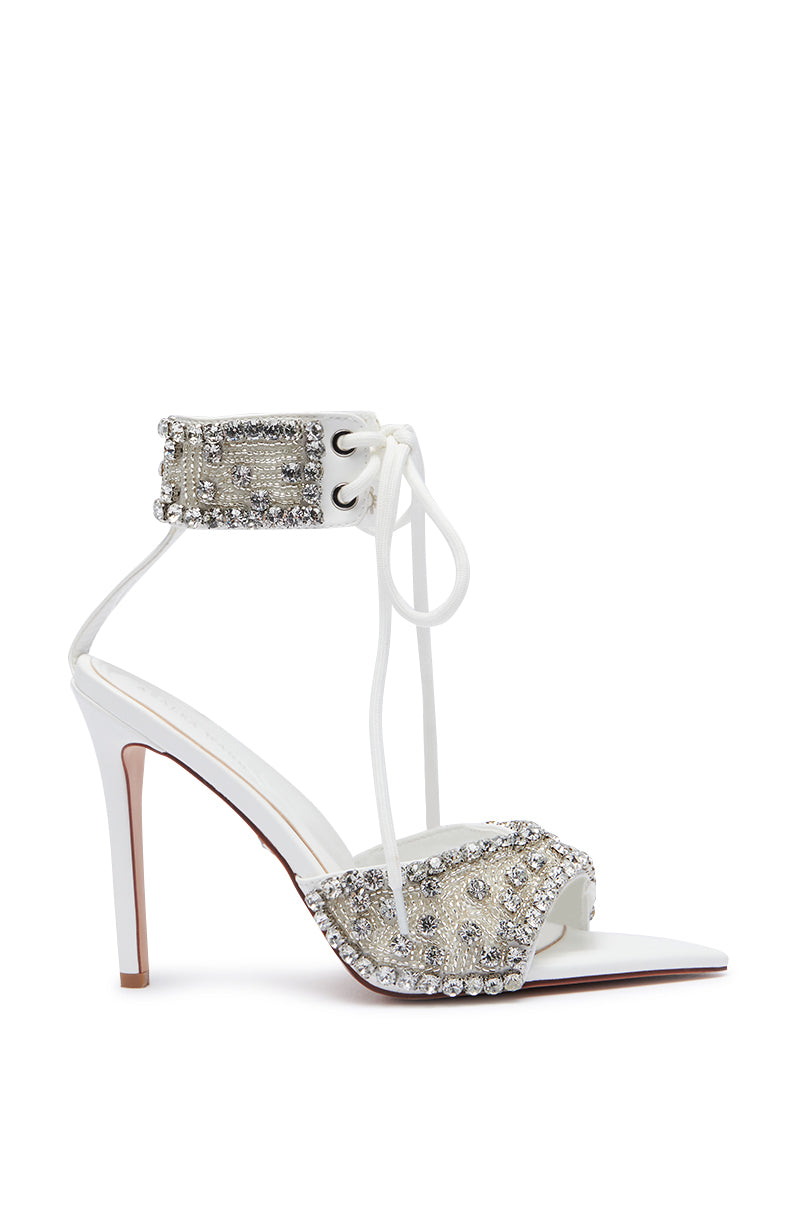 The AZALEA WANG ZELANIA WHITE SANDAL is a stunning white faux leather sandal embellished with shimmering silver rhinestones. It features an ankle strap that laces up and ties in the front, and the toe strap is also decorated with rhinestones. The sandal showcases a slim stiletto heel, a slightly pointed open toe, and includes a comfort cushion insole for added ease.