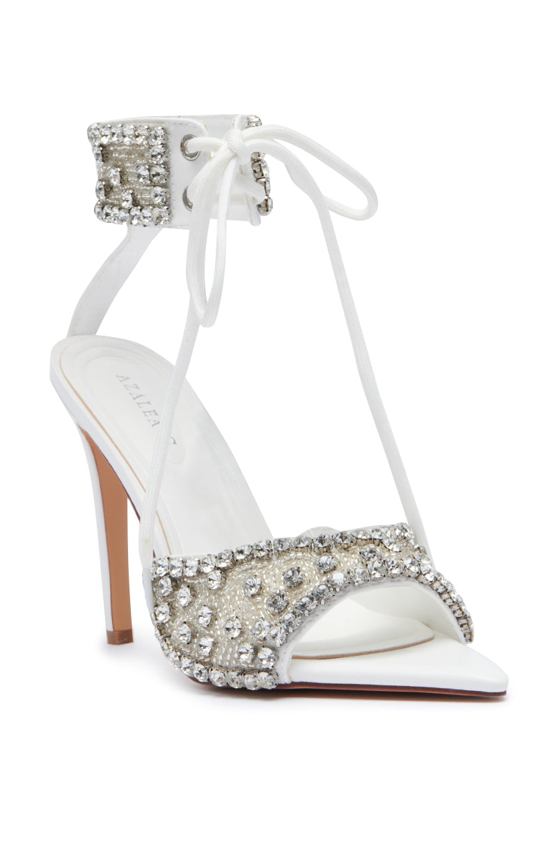 The AZALEA WANG ZELANIA WHITE SANDAL is a high-heeled, faux leather stiletto with white laces that elegantly wrap around the ankle. The front strap and ankle area are beautifully adorned with silver sequins and sparkling rhinestones. This stylish shoe features a comfort cushion insole, a white sole, and an open toe design.