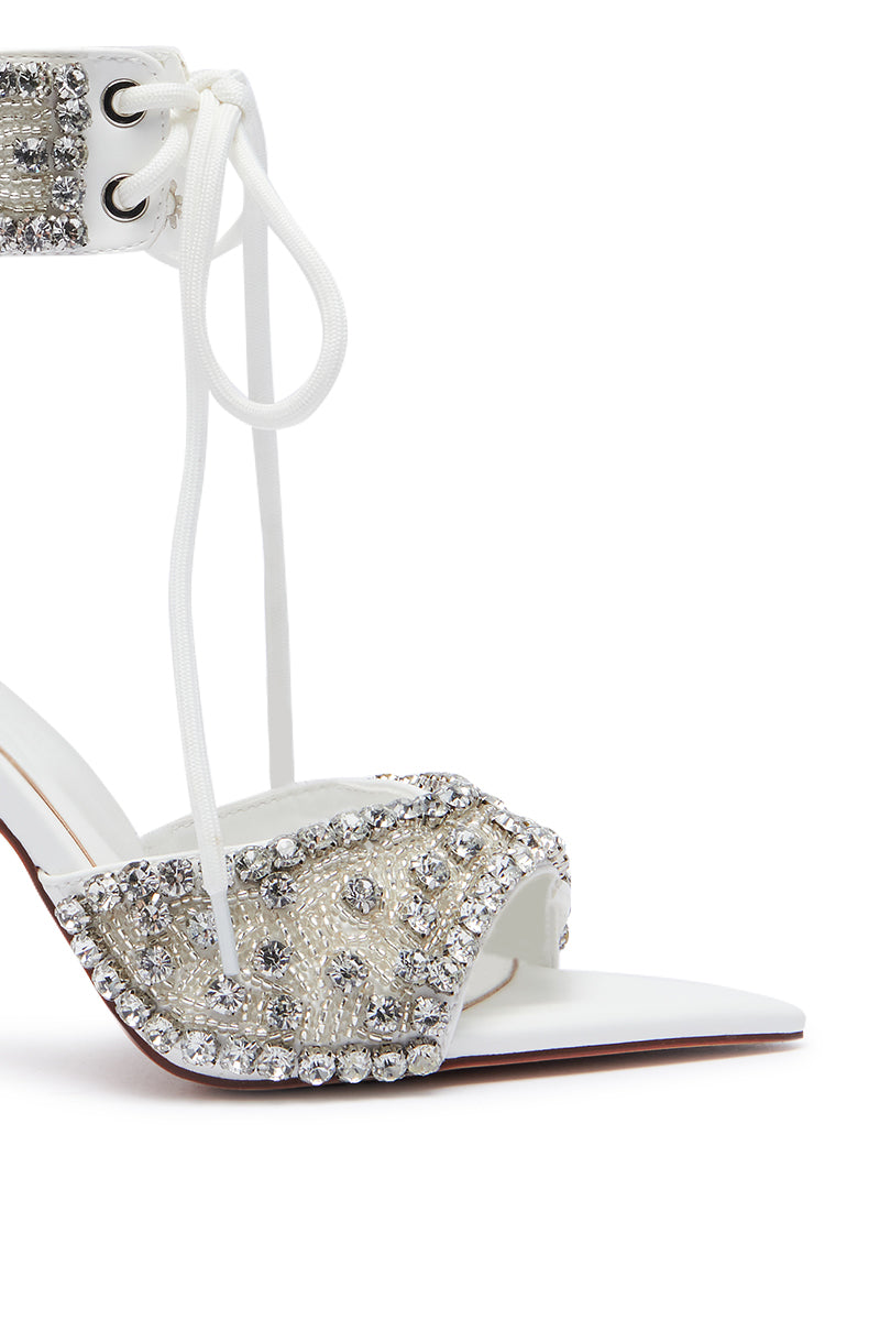 A close-up of the AZALEA WANG ZELANIA WHITE SANDAL, an elegant faux leather shoe adorned with sparkling rhinestones. The sandal features lace-up ankle straps, a pointed toe design, and a stiletto heel. Rhinestones decorate the straps and front, adding a touch of glamour to the footwear.