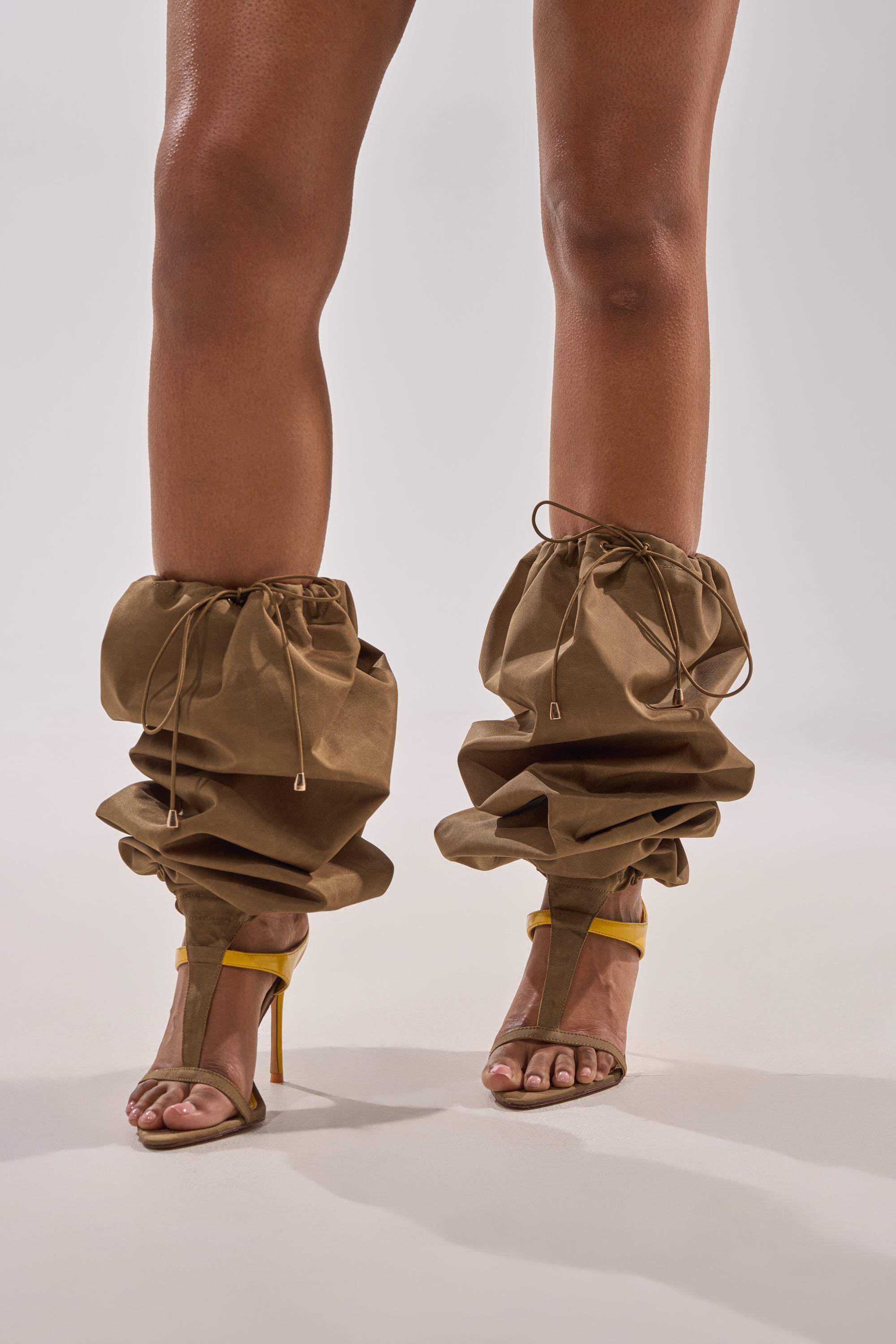 Someone wears the AKIRA GOLD ZIDANE OLIVE CANVAS SLOUCHY STILETTO SANDAL with loose, ruched brown leg coverings tied with drawstrings from ankle to lower calves. The plain, light background adds a subtle streetwear vibe. 1