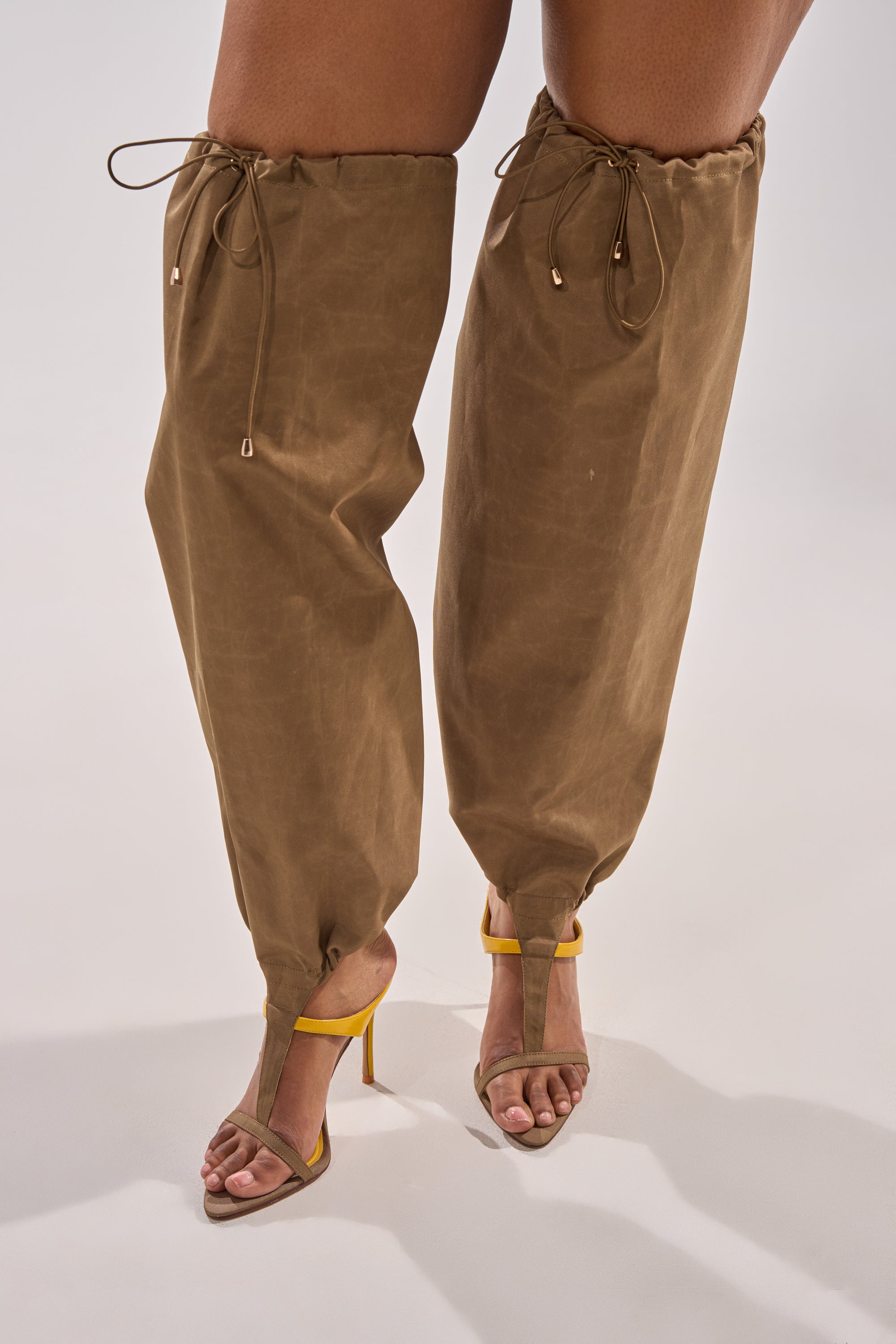 Wearing oversized, loose brown pants cinched at the knees and ankles, a person pairs them with AKIRA GOLD ZIDANE OLIVE CANVAS SLOUCHY STILETTO SANDALS for a bold, streetwear-inspired look on a plain background. 5