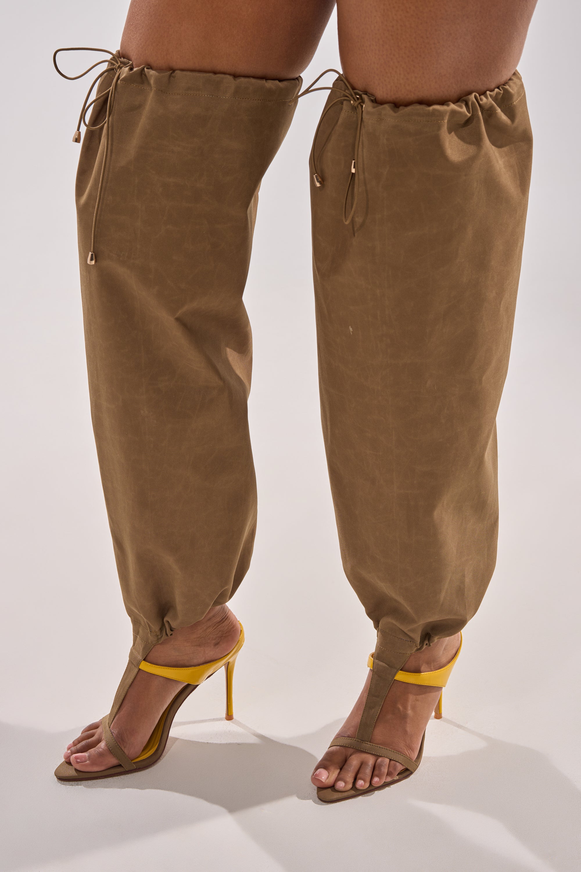 A person sports brown, loose drawstring pants with AKIRA GOLD ZIDANE OLIVE CANVAS SLOUCHY STILETTO SANDALS. The shot highlights their legs and feet against a plain light backdrop, showcasing modern streetwear flair. 6