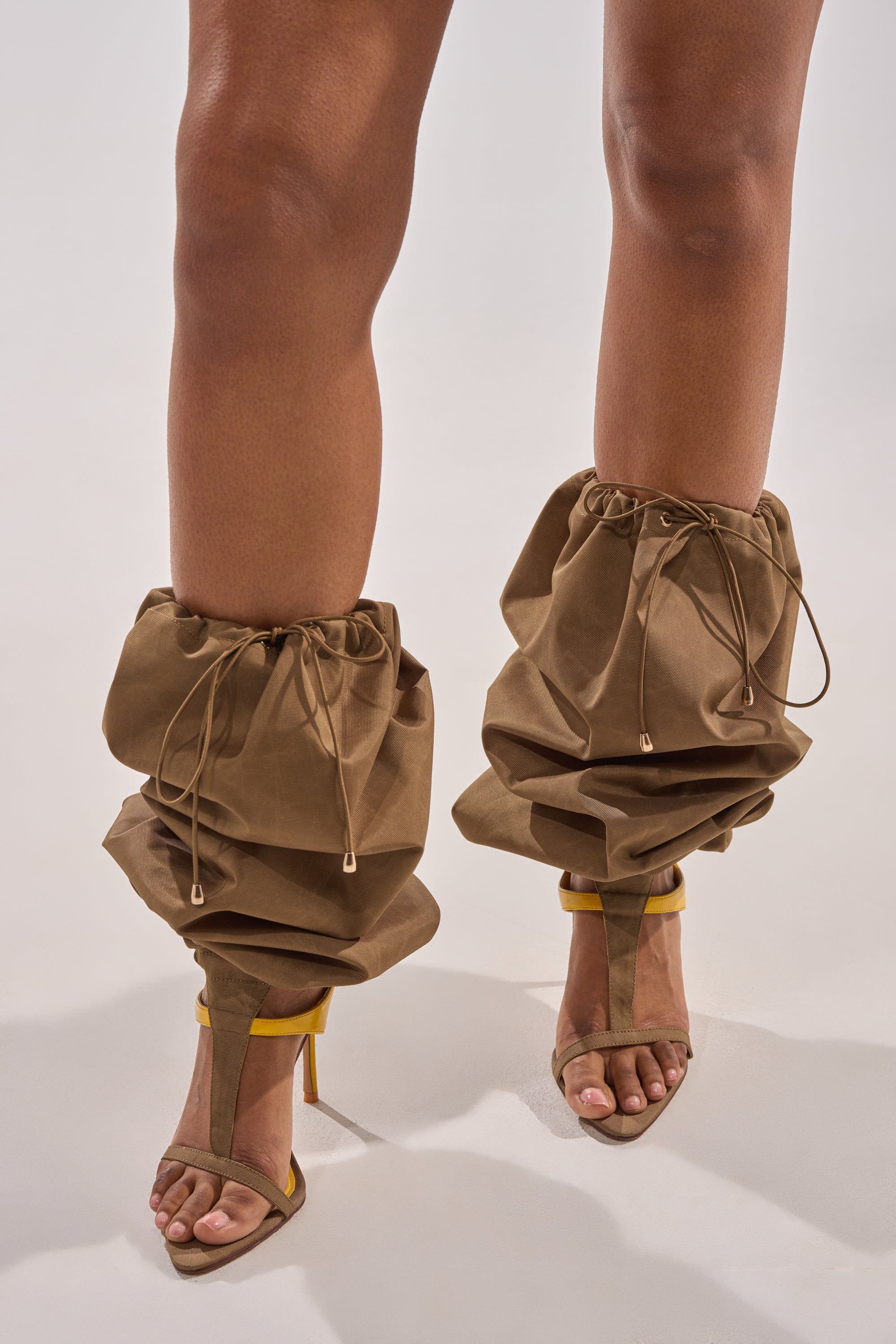 A person wears the AKIRA Gold Zidane Olive Canvas Slouchy Stiletto Sandal, featuring ruched brown fabric with drawstrings and yellow straps, against a plain light background. 3