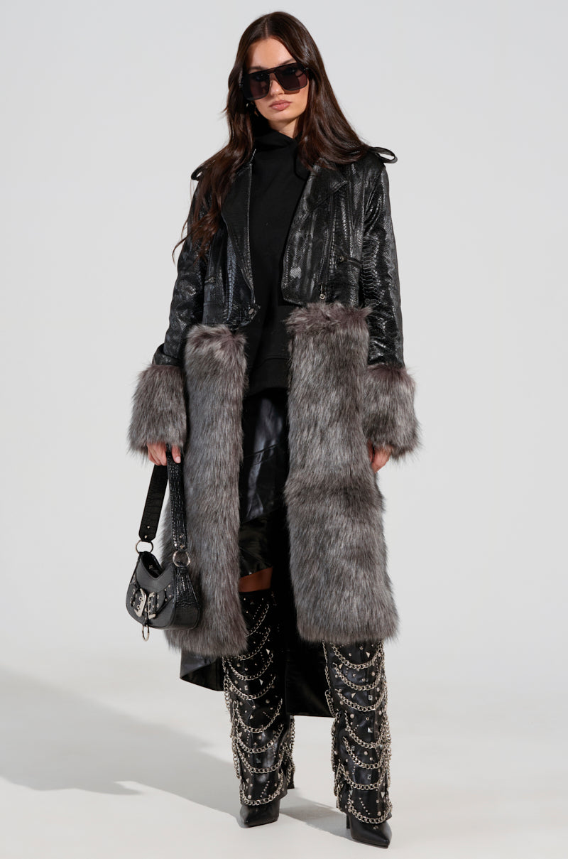 A person stands confidently, dressed in the 2 IN 1 MOTO FUR TRENCH and sleek black leather pants. They hold a small handbag, wear sunglasses, and pair knee-high boots adorned with chains. The croc-embossed vegan leather provides an edgy touch against the plain backdrop.
