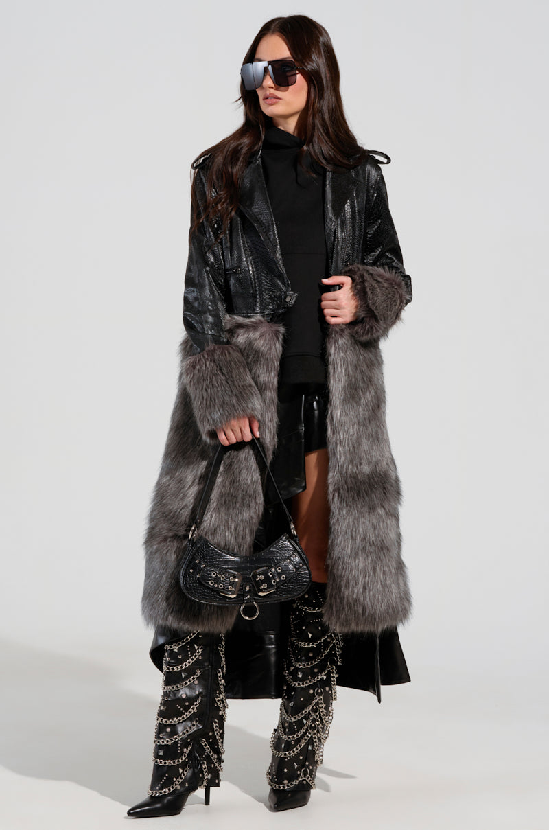 A woman stands confidently wearing a black outfit and a 2 IN 1 MOTO FUR TRENCH coat. She accessorizes with oversized sunglasses, a small black handbag, and knee-high boots with chain details, exuding an effortlessly chic, modern vibe.