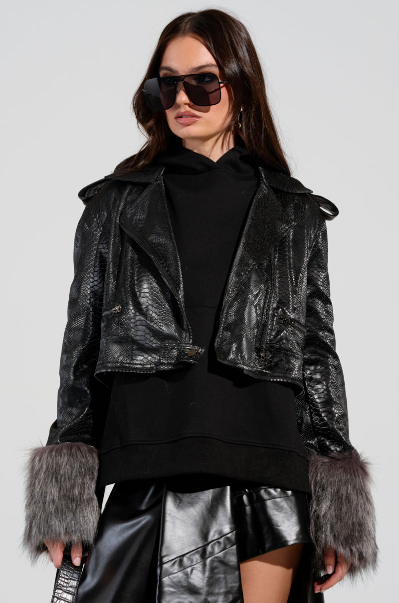 A person sporting large sunglasses and a 2 IN 1 MOTO FUR TRENCH, crafted from croc-embossed vegan leather with chic gray faux fur cuffs, exudes an effortlessly stylish vibe. Underneath the trench, they layer a black hoodie paired with a black skirt featuring an asymmetrical hem—strikingly fashionable against the plain background.