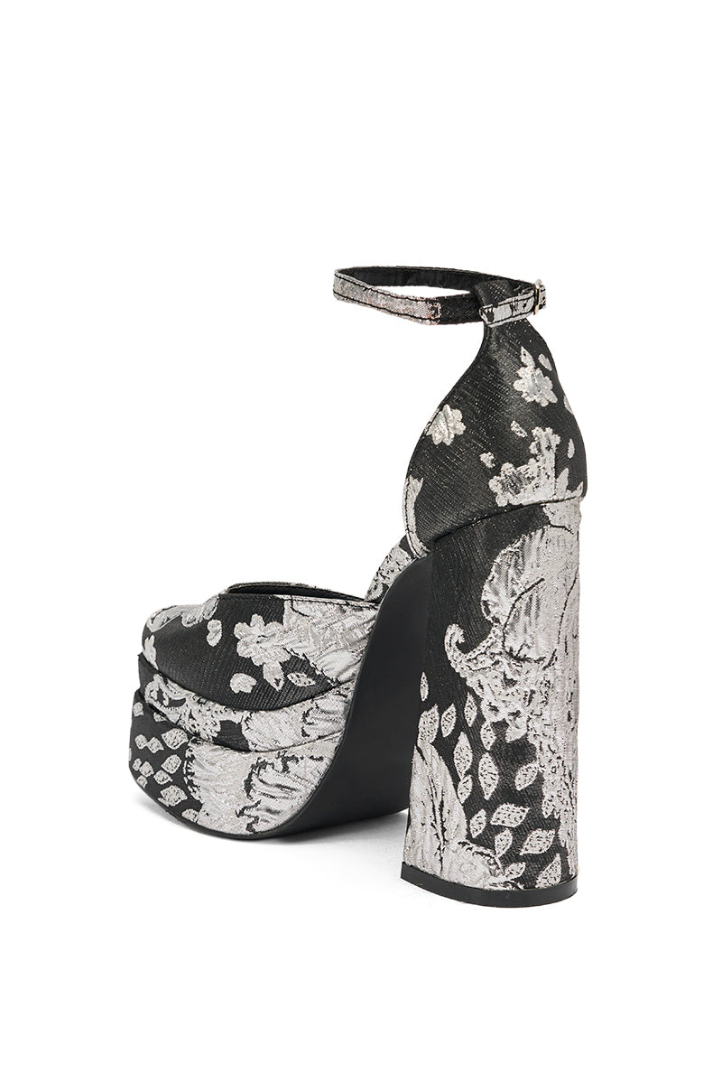 The AZALEA WANG ZIPPORAH BLACK BROCADE PUMP is a high-heeled platform sandal with a chunky heel, ankle strap, and bold black base adorned with silver floral and leaf embroidery in a textured brocade design.
