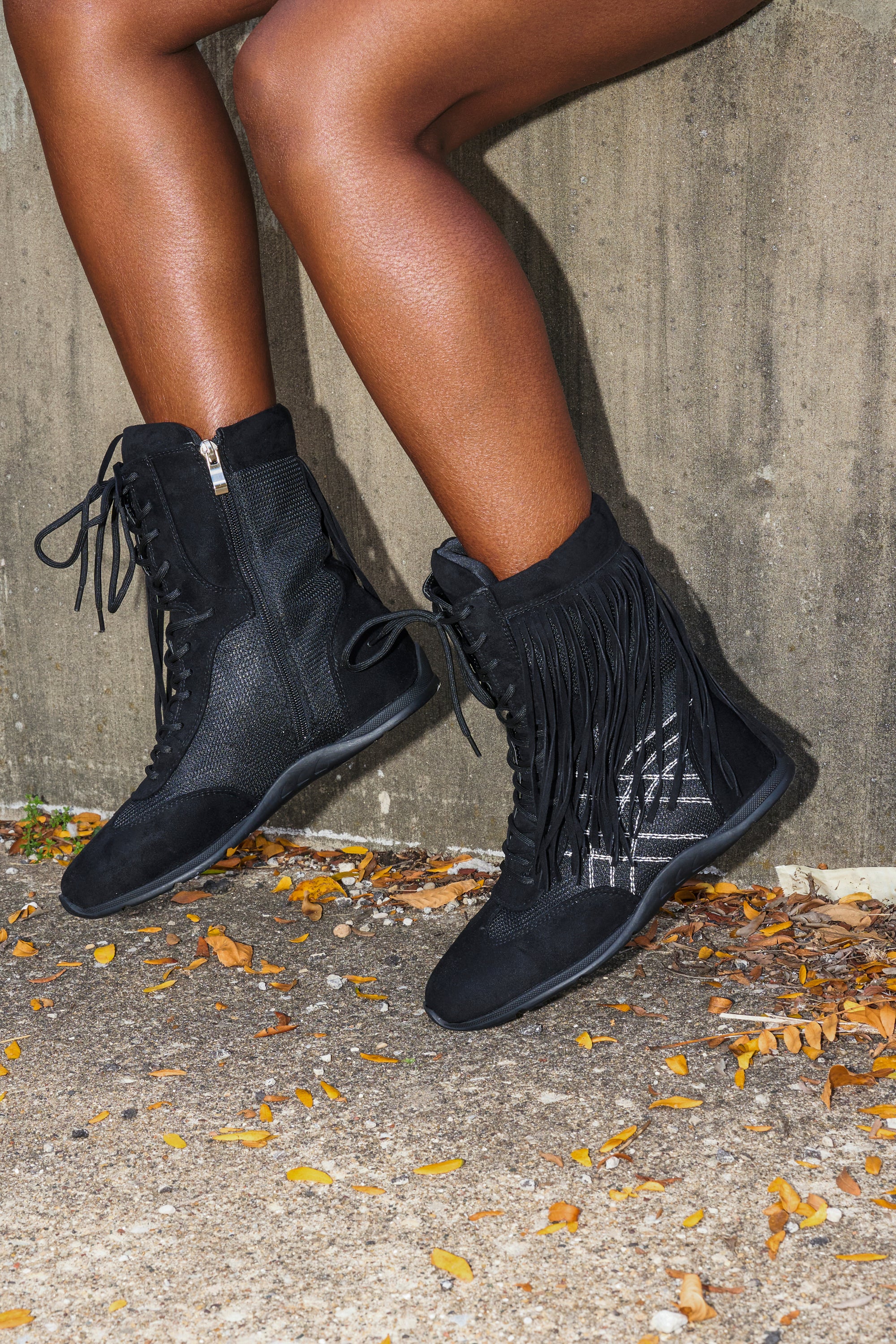 Wearing the AZALEA WANG ZITHER BLACK FLAT FRINGE BOOTIE, a person with legs crossed sits near a concrete wall amid scattered yellow leaves.