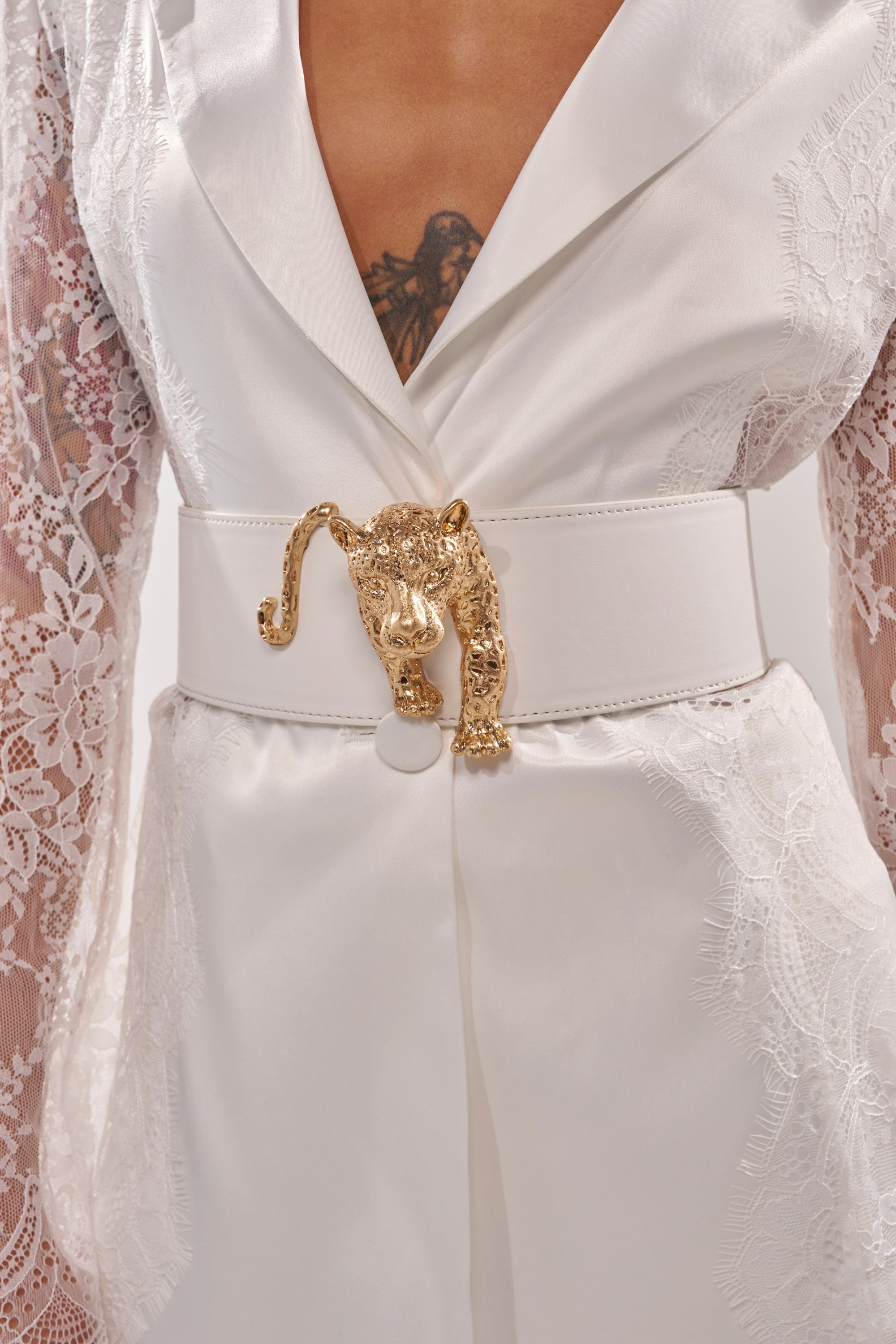 A person wearing a white lace-sleeved outfit with a plunging neckline, styled with the NO BOUNDARIES PU STRETCH BELT IN WHITE featuring a large, ornate gold panther head buckle at the waist. 0