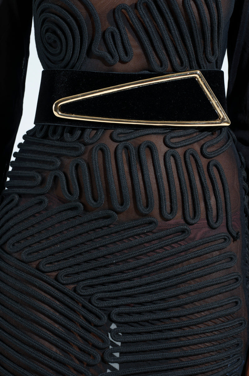Close-up of a sheer black dress with intricate swirling embroidery and luxurious velvet detailing. The GLORY ASYMMETRICAL BELT, featuring a large triangular gold buckle, cinches the waist to create a bold statement. 1