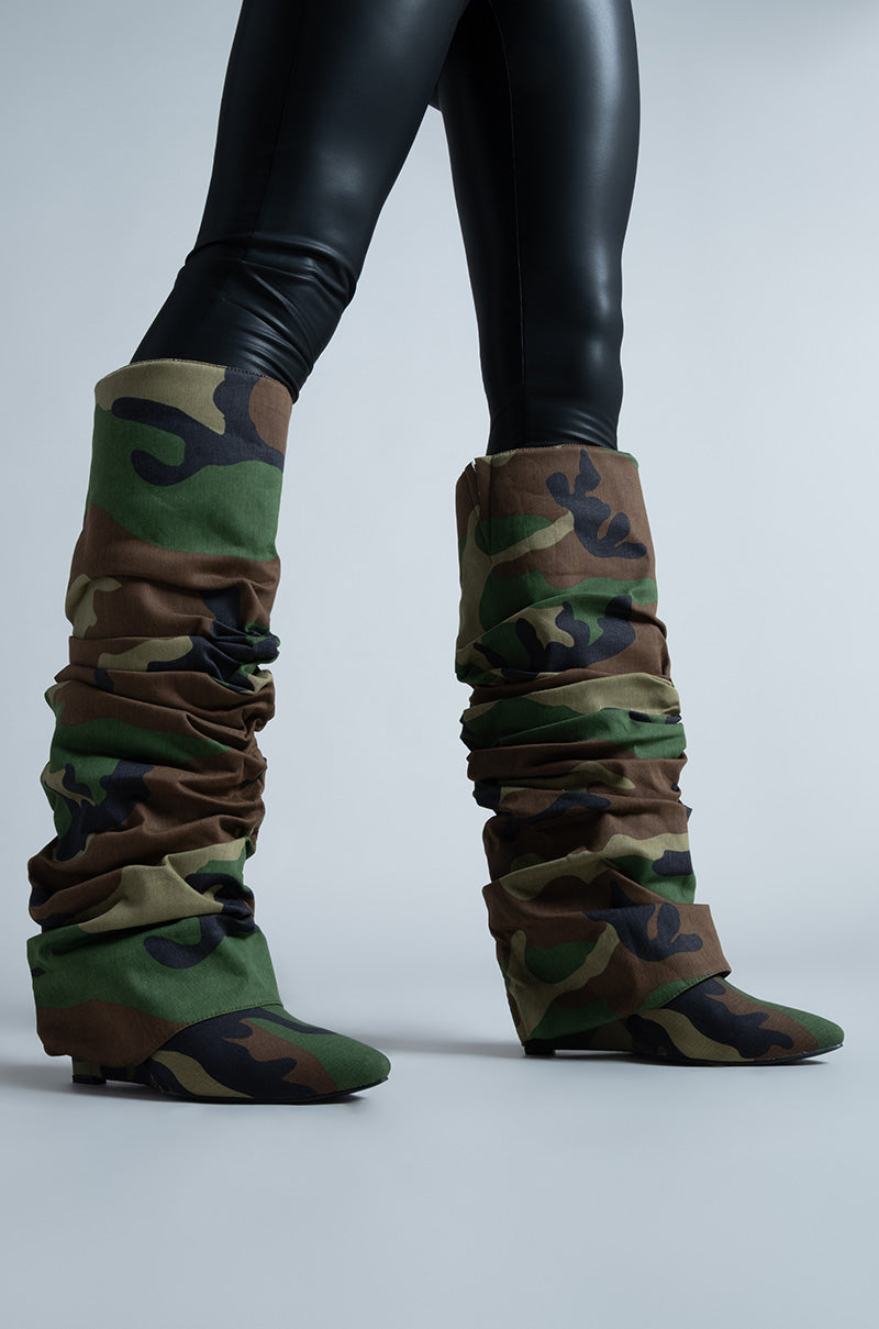 A person in black leggings wears AZALEA WANG ZUMMA CAMO WEDGE BOOTS, featuring a slouchy design and pointed toe, against a plain gray background.