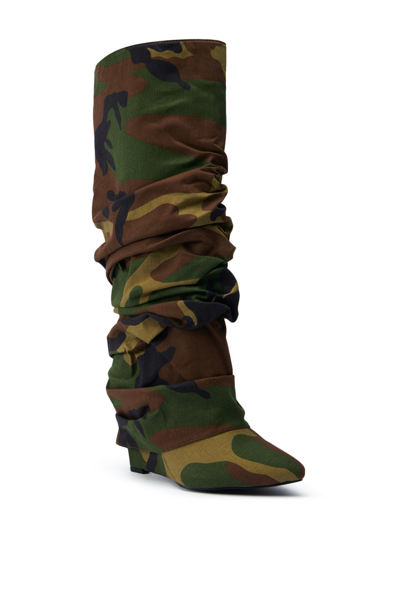 The AZALEA WANG ZUMMA CAMO WEDGE BOOT is a knee-high, slouchy boot with shades of green, brown, and black camo. It features a low wedge heel and pointed toe, set against a plain white background.
