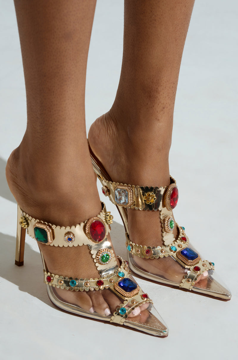 A close-up of a person wearing the AZALEA WANG ZYA MULTI GOLD EMBELLISHED PVC PUMP, which features gold straps with multicolored gems and transparent material at the pointed toe, contrasting beautifully with their dark skin against a plain white background.