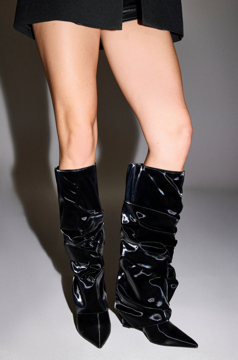 A person wearing a black skirt and AZALEA WANG ZYLLA BLACK PATENT WEDGE RUCHED BOOTS with pointed toes stands on a light gray surface.