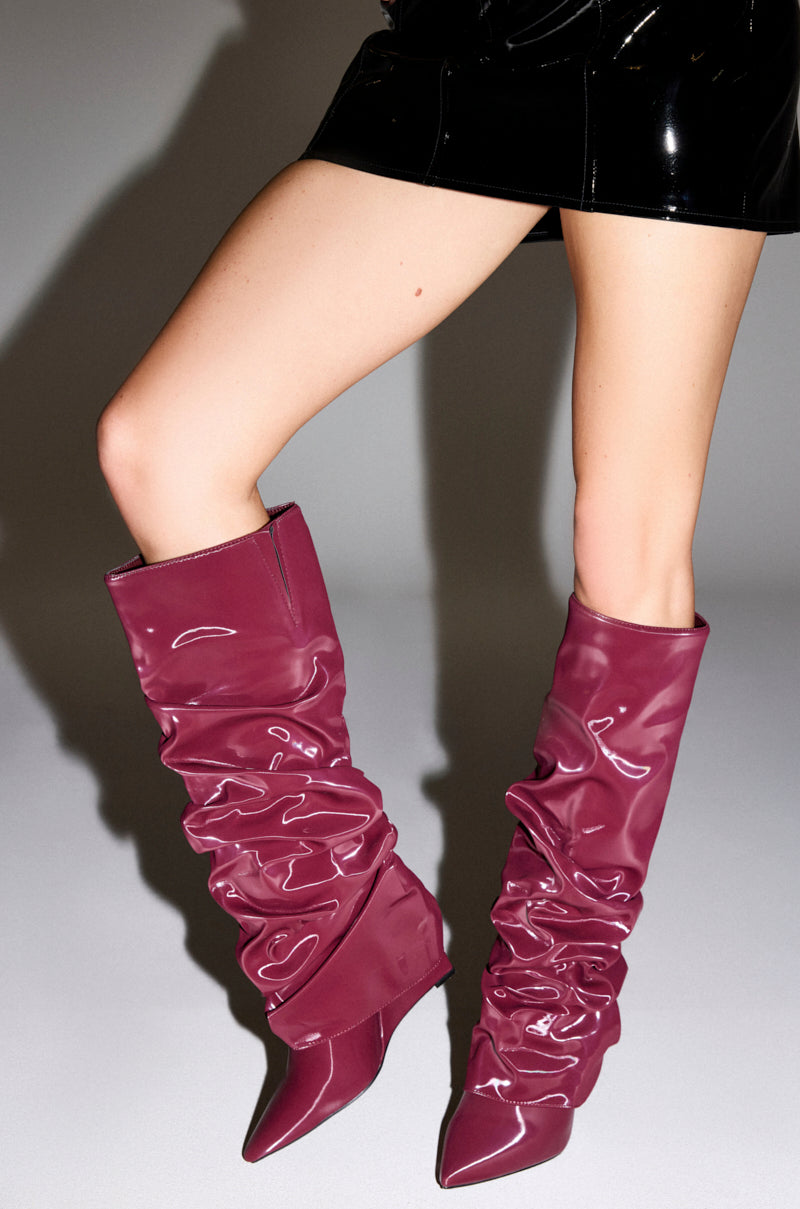 A person wears a shiny black mini skirt and AZALEA WANG ZYLLA BURGUNDY PATENT WEDGE RUCHED BOOTS, featuring a slouchy fold-over design, standing against a plain light background.