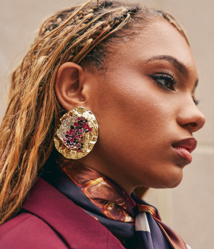 A close-up side profile of a woman wearing a large, textured gold earring embellished with red and pink gemstones, paired with a burgundy blazer and patterned scarf.