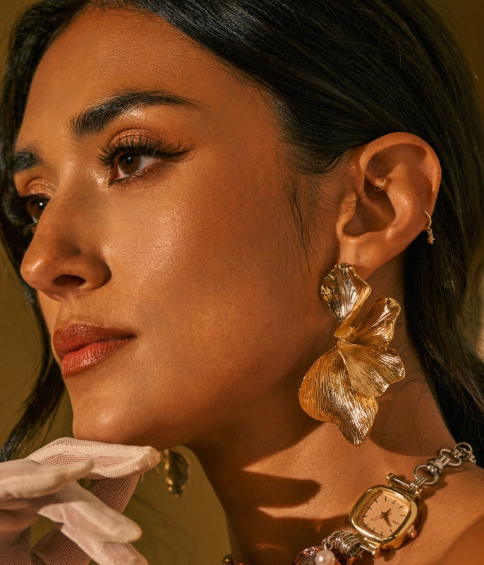 A close-up portrait of a woman wearing large textured gold leaf drop earrings, a gold watch with a chain strap, and a white glove, set against a warm, solid background.