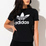 A woman with long dark hair is wearing an ADIDAS WOMENS TREFOIL TEE, featuring the brand's logo in white. She is standing against a plain white background, gazing slightly to the right with one hand resting by her side.