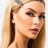 A woman with long, straight blonde hair is shown in a close-up shot, wearing the AKIRA BOBBI PIN adorned with jewels on the side. She has winged eyeliner, long eyelashes, and pink lipstick, showcasing a subtle and elegant makeup look against a plain, light background.