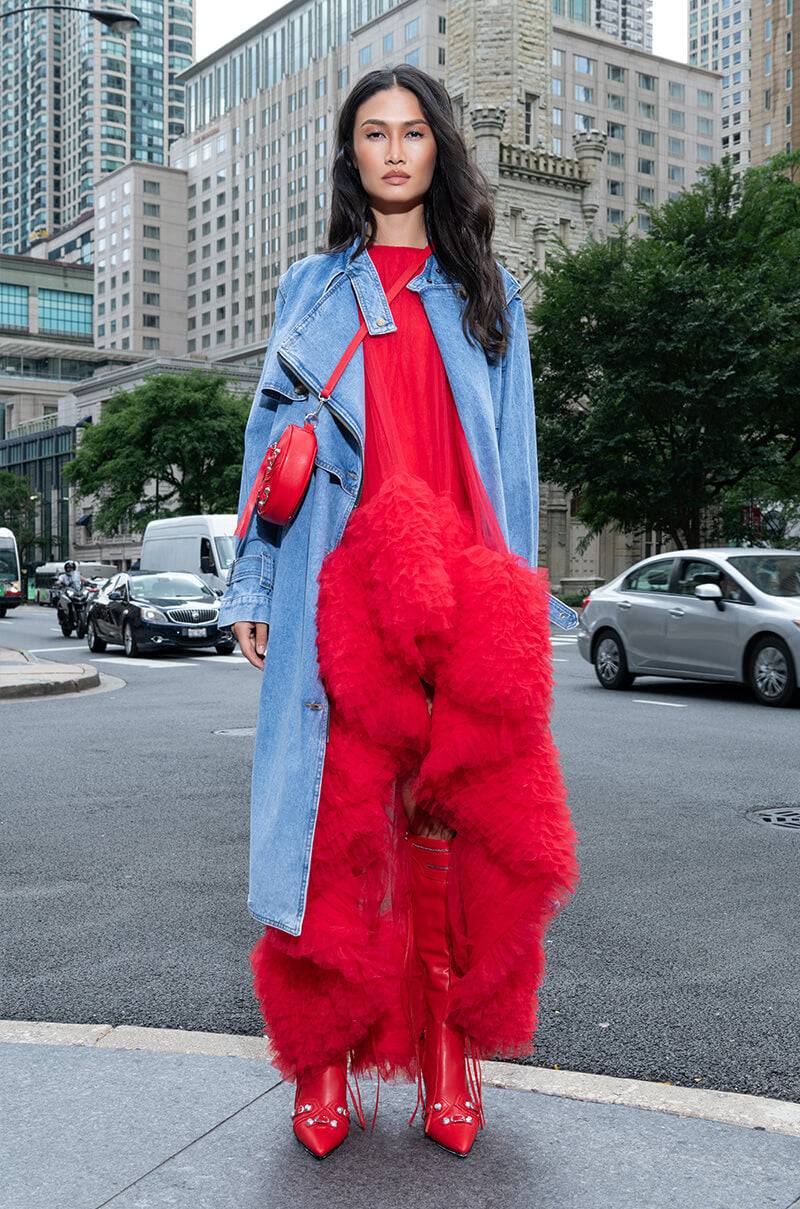 A woman stands confidently on a city street wearing the ARI TULLE RUFFLE HIGH LOW MINI DRESS IN RED paired with a denim jacket and matching boots. Her red purse completes the ensemble against an urban backdrop of buildings and cars.