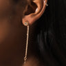 Close-up of a person's ear adorned with multiple earrings. They are wearing the ARTEMIS EAR CUFF SET, which features a small, round gold stud earring with a long, thin, square-link chain hanging down, paired with a small hoop earring on the cartilage. The dark background emphasizes the jewelry.