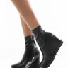 A close-up image shows a person wearing the stylish AZALEA WANG CAN'T BREAK US WEDGE BOOTIE IN BLACK. The boots feature a side zipper and a textured sole. The person's legs are slightly bent, set against a clean, bright white background.