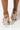 Close-up of a person wearing the AZALEA WANG LEFT YOUR SOUL BY THE RIVER HEEL SANDAL IN SILVER, featuring transparent straps, thin clear stiletto heels, and decorative shiny elements. The photo focuses on the back of these uniquely designed sandals, highlighting the person's ankles and heels.