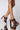 A close-up of a person's legs wearing the AZALEA WANG LET'S SLOW IT DOWN CHUNKY SANDAL IN CHOCOLATE, showcasing the brown high-heeled sandals with a stylish braided strap design. The sandals feature block heels, and the person's legs are crossed at the ankle, standing on a reflective surface against a plain background.