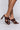 A person is wearing the AZALEA WANG LET'S SLOW IT DOWN CHUNKY SANDAL IN CHOCOLATE, which features dark brown, high-heeled sandals with a braided strap design. The shoes are open-toed and have a chunky heel. The background is plain and white.