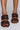 Close-up of a pair of feet wearing the AZALEA WANG LET'S SLOW IT DOWN CHUNKY SANDAL IN CHOCOLATE. The sandals feature thick braided straps over the toes and insteps, complementing the neatly painted pink toenails of the wearer. The background is a neutral gray.