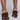 A person wearing the AZALEA WANG LET'S SLOW IT DOWN CHUNKY SANDAL IN CHOCOLATE stands against a light gray background. The image captures only the lower legs and back view of the shoes, emphasizing their intricate braided detail and sturdy heel design.