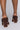 A person wearing the AZALEA WANG LET'S SLOW IT DOWN CHUNKY SANDAL IN CHOCOLATE stands against a light gray background. The image captures only the lower legs and back view of the shoes, emphasizing their intricate braided detail and sturdy heel design.