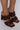 A close-up of a person's feet wearing the AZALEA WANG LET'S SLOW IT DOWN CHUNKY SANDAL IN CHOCOLATE, showcasing high-heeled sandals with thick, woven, dark brown leather straps. The sandals feature an open toe design and are paired with pink nail polish on the toes. The heels are block-style and square-shaped.