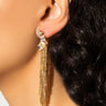 Close-up of a woman wearing the BABY DRIVER ELEGANT EARRING, a long, gold chandelier earring featuring multiple dangling strands. Her dark, wavy hair frames the scene, with a blurred background that enhances the prominence of the elegant and sparkly earring, suggesting a formal or glamorous style.