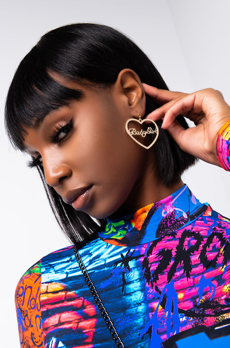 A person with short black hair wears a colorful, graffiti-patterned shirt. They hold up a BABY GIRL HEART HOOP EARRING, featuring a gold heart shape with the word "BabyGirl" in the center. The background is white and minimalistic.