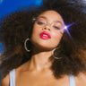 A woman with voluminous curly hair poses against a bright blue sky with clouds. She is wearing large hoop earrings and stylish rectangular BACK TO BACK SUNNIES that reflect a glare of light. Her makeup includes vibrant red lipstick and subtle eye shadow.