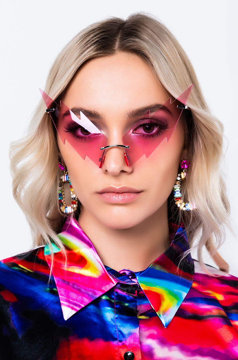 A woman with blonde hair wearing angular pink BAT WING SUNNIES and large, colorful, beaded hoop earrings. She has pink eye shadow and a colorful, abstract-patterned shirt with a mix of red, blue, yellow, and green hues. The background is plain white.
