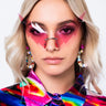 A woman with blonde hair wearing angular pink BAT WING SUNNIES and large, colorful, beaded hoop earrings. She has pink eye shadow and a colorful, abstract-patterned shirt with a mix of red, blue, yellow, and green hues. The background is plain white.